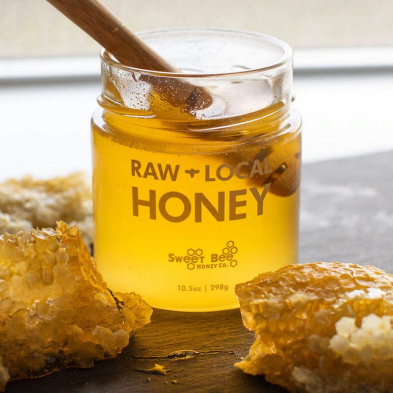 Jar of raw local honey with a wooden spoon and honeycomb on a wooden surface.